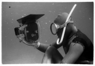 Black and white side angle shot of a snorkeller sitting on the seafloor holding an underwater camera 