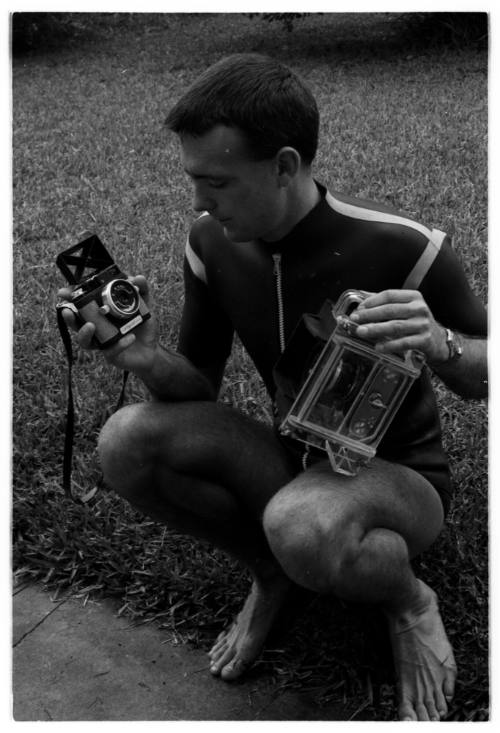 Black and white photo of Ron Taylor crouched while holding two cameras