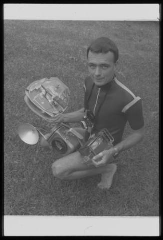 Black and white photo of Ron Taylor crouched while carrying four of his cameras