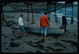 Three people standing around a small dinghy on a beach