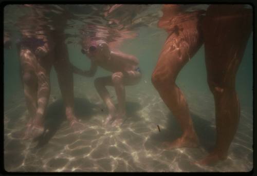 Underwater shot of shallow sandy sea floor showing legs of three children and two adults, one child crouched looking underwater with goggles on