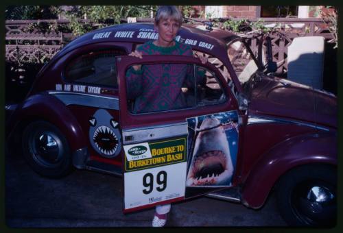 Sideview shot of Valerie Taylor standing with maroon Volkswagen Beetle with shark-related stickers on it, including cartoon shark jaw on front hood