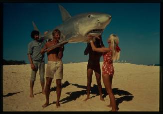 Shot of Valerie Taylor and group of people holding up a large prop shark on beach sand