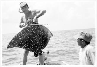 Shot of freediver holding a caught Spotted Eagle Ray standing on board a boat sea with a second passenger in foreground