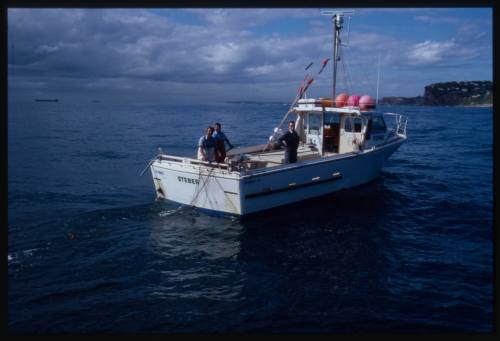 Fishing boat at sea with three people on board and net trailing off stern