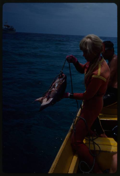 Valerie Taylor standing on small boat deck holding bait on a line, Ron Taylor behind her also on deck