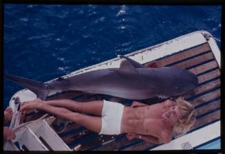 Person laying down on deck of boat with a similar sized Silver Tip Shark out of water laying beside them