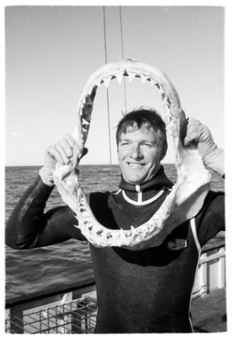 Black and white  Topside shot person in wetsuit holding up a shark jaw on deck of a ship at sea