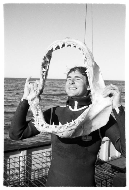 Black and white  Topside shot person in wetsuit laughing holding up a shark jaw on deck of a ship at sea