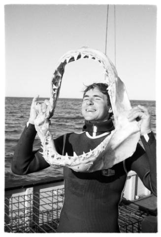 Black and white  Topside shot person in wetsuit laughing holding up a shark jaw on deck of a ship at sea