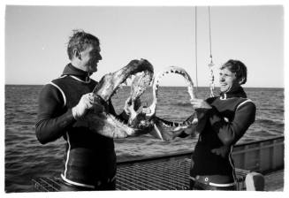 Black and white  Topside shot of two people in wetsuits laughing holding up a shark jaw each on deck of a ship at sea