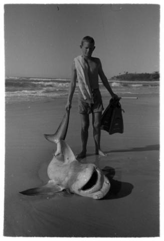 Black and white  Shot of person standing at water's edge holding the tip of a caught shark's tail that is laying on sand