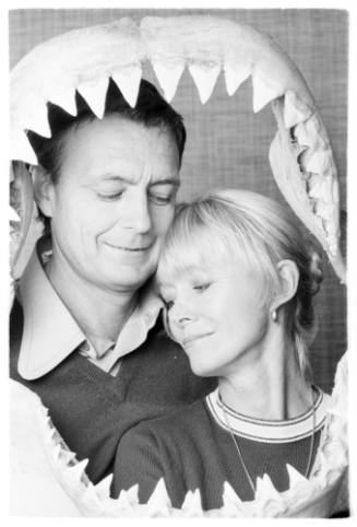 Black and white  Portrait shot of Ron and Valerie Taylor with eyes closed framed by a shark jaw Ron is holding up