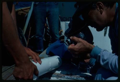 Person looking through camera at cylinder with bite marks that a second person is holding, on deck on board a boat