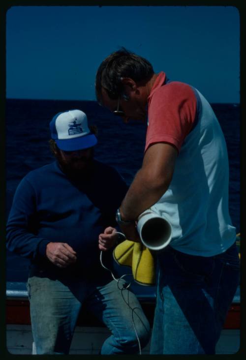 Ron Taylor holding a cylindrical pipe and second person holding a wire on board a boat