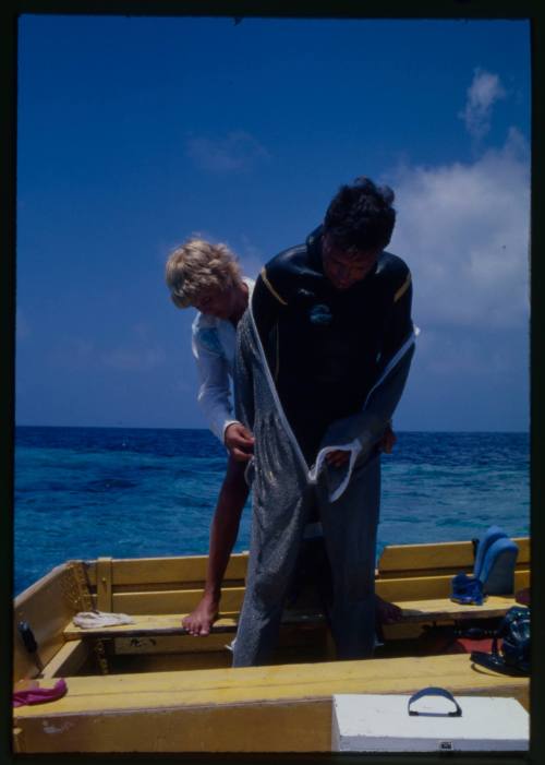 Valerie Taylor assisting Ron Taylor into full mesh suit standing on board a boat