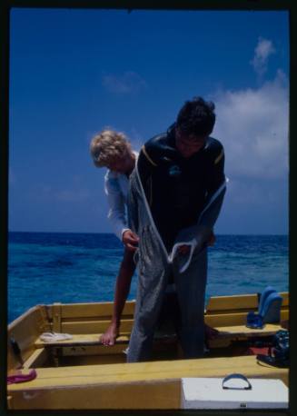 Valerie Taylor assisting Ron Taylor into full mesh suit standing on board a boat