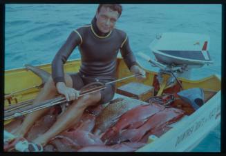 Shot of Ron Taylor on dinghy holding spearfishing gun, sitting with catch of fish laid out on seats and deck