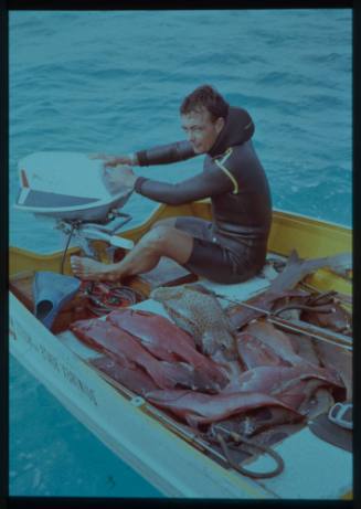 Shot of person on dinghy operating the engine, sitting with a catch of fish and spearfishing guns laid out on seats and deck