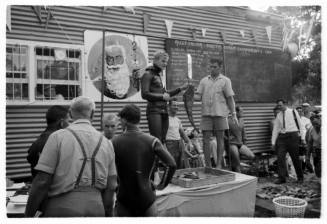 Person holding caught fish near weighing hook by whiteboards with groups of people around