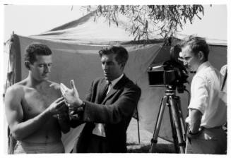 Medium shot of three people standing by filming equipment with a large tent pitched in background