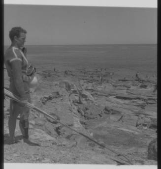 Shot of Ron Taylor in dive gear holding spear rod standing on rock platform