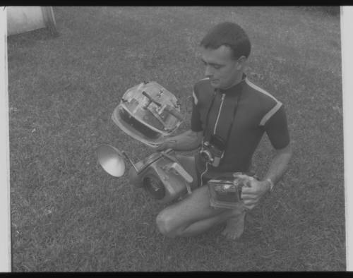 Shot of Ron Taylor in wetsuit holding underwater camera equipment while kneeling on grass