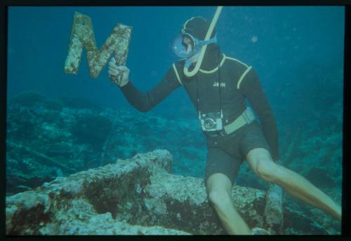 Underwater shot at seafloor of freediver holding a colonised and encrusted letter 'M'