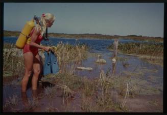 Shot of Valerie Taylor standing in ankle deep water with scuba gear amongst grasses near water's edge