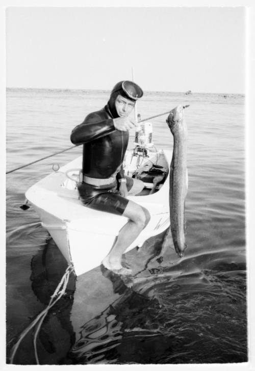 Black and white shot of diver sitting on bow of dinghy holding up a caught eel speared through the head