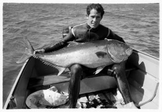 Black and white topside shot of diver sitting at back of dinghy holding a large Trevally on their lap