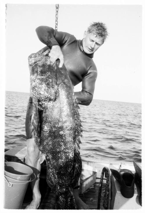 Black and white topside shot of diver holding up an Atlantic Goliath Grouper standing on a dinghy at sea