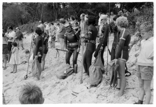 Black and white shot of large group of people with group in dive gear holding spearfishing rods at forefront of group standing on rocky land