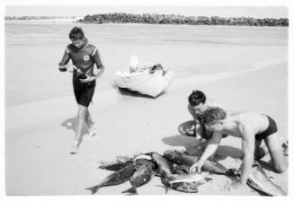 Black and white shot of diver and two people around a haul of caught fish on the ground with dinghy in midground at the water's edge