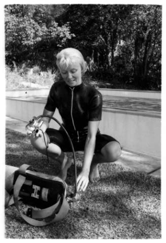 Black and white shot of Valerie Taylor in wetsuit holding regulator crouched beside air tank laying on ground
