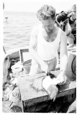 Black and white shot of person cutting into caught fish on board a dinghy at sea with divers suiting up in righthand background