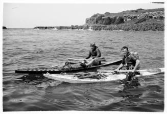 Black and white shot of two people in kayaks at sea with Cook Island shoreline in background