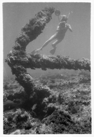 Black and white underwater shot of large encrusted anchor on rocky seafloor with diver in background