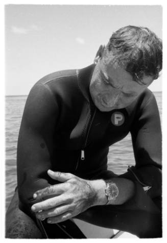 Black and white shot of diver Peter Woodhead in wetsuit sitting with sea in background
