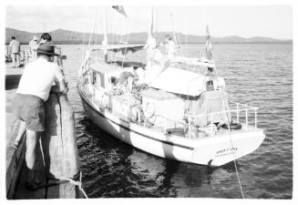 Black and white shot of yacht docked at wharf with onlooker standing on wharf edge in lefthand foreground