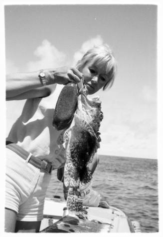 Black and white shot of Valerie Taylor holding a caught fish by a hook in its lip at the edge of a boat at sea