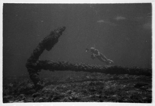 Underwater shot SIRIUS anchor laying horizontal on rocky seafloor with diver swimming beside in background