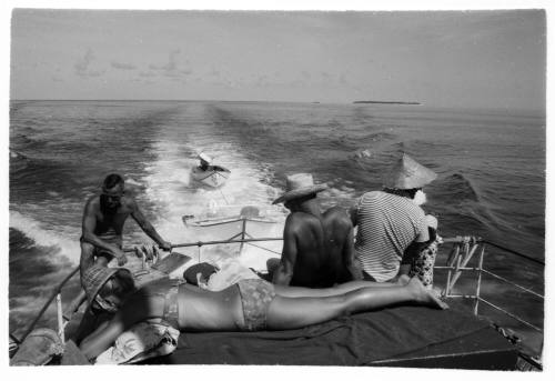 Black and white topside shot of group sitting at the stern of a boat at sea with two dinghies trailling behind