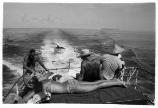 Black and white topside shot of group sitting at the stern of a boat at sea with two dinghies trailling behind