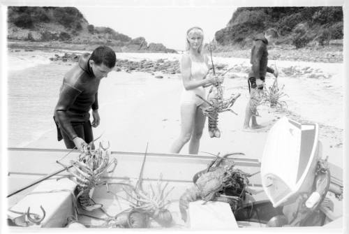 Black and white shot of three divers holding large caught crayfish by a dinghy on shore at the waters edge