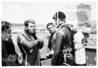 Black and white shot of four divers with one fully suited up with flag in background