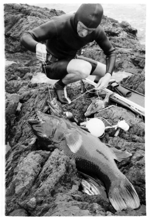 Black and white shot of freediver squatting on rock platform with caught fish on floor