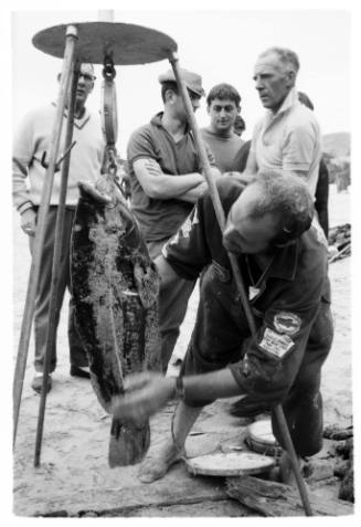 Black and white shot of person weighing fish on scale with group of people in background