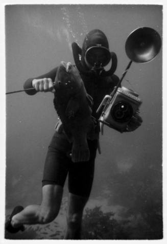 Black and white underwater shot of freediver near seafloor holding camera equipment and caught fish pierced with spear rod