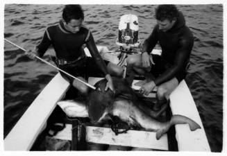 Black and white topside shot of two divers sitting at stern of dinghy, one holding a spear rod that is piercing a caught fish